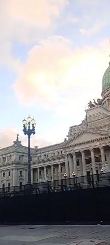 The video shows the Argentine National Congress building in Buenos Aires, Argentina. The camera pans from the left side of the building to the right, displaying the large green dome, numerous columns, and decorative statues.  A man wearing a blue jacket and white cap walks in front of a black fence in the foreground. Several lampposts are visible along the building's facade. The sky in the background is pale with a few clouds, suggesting either dawn or dusk.  The sound of bells can be heard.