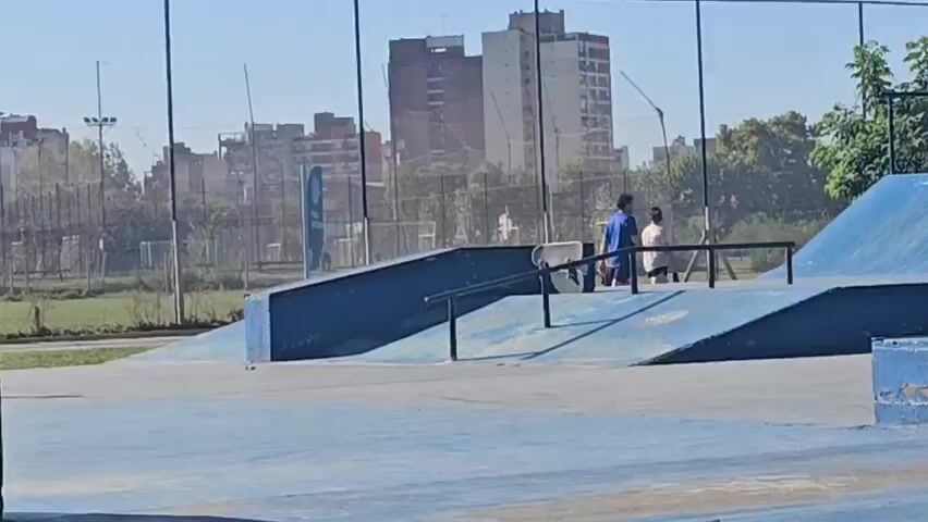The video shows a male skateboarder performing a trick in a skate park. He is shirtless, wearing patterned shorts, and riding on a blue concrete surface. The trick involves him jumping and getting his board over a black metal rail. Behind him, there are other people in the skate park, including a few observers. The backdrop includes buildings, trees, and a metal fence. The lighting indicates it is a sunny day. The skate park features blue ramps and other obstacles typical of such a facility.