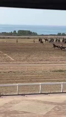 Several people are riding horses on a dirt track in a race.  The track is surrounded by a white fence, and a small group of people are standing in the distance.  Behind the track, there is a body of water and some trees in the distance. The video is being taken from a building looking out onto the track.  People in the crowd are heard cheering loudly.