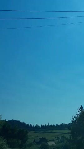 The video shows a landscape with a clear blue sky and a line of trees in the distance. There are some houses and fields visible between the trees and the foreground. A few power lines and poles are also present in the scene. The overall impression is of a rural or semi-rural setting on a sunny day.