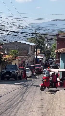 The video depicts a street scene in a town with a slight incline. There are various vehicles on the street, including cars, a truck, motorcycles, and several red auto rickshaws or tuk-tuks. One of the tuk-tuks is idling on the side of the road. The street is lined with buildings, some appearing older with red-tiled roofs, and a newer one with concrete block construction. Overhead, there is a dense network of power lines. People are walking down the street. In the background, there is a mountain range under a blue sky with scattered clouds.