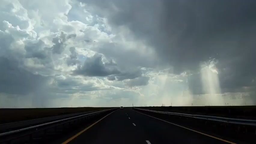 The video shows a point-of-view shot from inside a vehicle traveling on a highway. The sky is dramatic with heavy, dark storm clouds interspersed with patches of bright sunlight breaking through. The highway is asphalt with a yellow line on the right edge and a white dashed line marking the center. Guardrails are visible on both sides of the road. In the distance, a white vehicle is visible on the highway. The landscape on either side of the road appears flat and barren, with some faint structures or power lines visible on the horizon. The lighting suggests a storm is either approaching or passing.