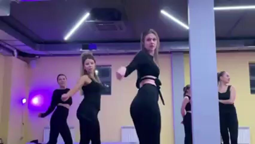 In a dance studio, a group of five women are practicing a dance routine. They are dressed in black workout attire, consisting of leggings and tops, some cropped to expose the midriff. The women are positioned in a line, executing synchronized movements that involve arm extensions, hip swivels, and bending forward. The studio has light yellow walls, a light grey ceiling with exposed ductwork, and bright white, angular light fixtures. There is a white pillar in the foreground, partially obstructing the view of the dancers. A small window with a dark frame can be seen in the background. The overall lighting in the studio is a mix of white and purple hues.