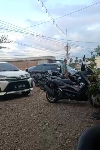A video shows a parking area with gravel, in front of a one story building. A white Toyota Avanza with license plate BL 1634 NM is parked on the left. To the right of it is a small tree in a square planter. Two black vans are parked behind the Avanza, and several black scooters are lined up near a small plant in the foreground. Overhead are electrical lines and a string of lights. A man wearing a green shirt and brown sarong walks in the background. The sky is overcast.