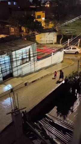 A night scene from an elevated angle captures two people walking on a wet, brick-paved street. The person in front is wearing a bright pink coat, while the other individual, possibly an adult, is clad in dark clothing.  The street is lined with simple, single-story buildings, some with corrugated metal roofs and whitewashed walls. Power lines crisscross overhead, illuminated by streetlights.  The perspective is from a rooftop, as evidenced by the corrugated metal and exposed rebar in the foreground.