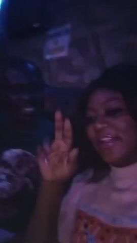 Two people, a man and a woman, are in a dimly lit indoor setting, possibly a club or bar, due to the low lighting and background music. The man is behind and to the left of the woman. Both are smiling. The man is wearing a dark green shirt and blue jeans. The woman is wearing a tan top with a graphic design on the front. A "No Smoking" sign is visible on the wall. The woman points at the camera.