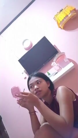 A young woman with dark hair is sitting on the floor, holding a pink compact mirror. She is looking into the mirror and appears to be applying something to her face, possibly makeup. She is wearing a dark purple sleeveless top. The background is a pink wall with a flat-screen television mounted above a white shelf. On the shelf are two pink containers. There is also a white clock on the wall above and to the left of the television. A yellow and gold decorative item is visible at the top of the image.