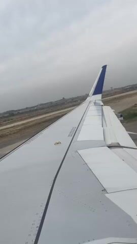 The video shows an aerial view from the wing of an airplane. The plane appears to be on the tarmac, possibly preparing for takeoff or just having landed. The wing is white with a blue tip. In the background, there is a runway, some buildings, and a cloudy sky. The camera angle is fixed, and the video is short, lasting only a few seconds. No people are visible in the video. The overall impression is one of travel and anticipation of flight.
