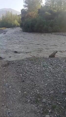This media file shows a natural scene with a motorbike. The scene is a riverside, with a body of water flowing gently. The water appears somewhat murky, suggesting it might be carrying sediment. The riverbanks are lined with rocks and gravel, and there is vegetation growing on the slopes leading up to the river. Trees and foliage are visible on the opposite bank and in the background. The sun is visible in the sky, casting a warm, hazy light, indicating it is likely late afternoon or early morning. The bright sunlight creates lens flare effects in the video.

The primary object in the foreground is a motorbike, parked on the rocky riverbank. It is a red and black motorcycle, appearing to be a dirt bike or dual-sport model. The motorbike is angled so that its side and rear are visible. It has a worn appearance, suggesting it has been used on rough terrain.

There are no people visible in the media. The activity taking place is simply the presence of the motorbike at the riverside, with the natural flow of the water. The setting is a rural, outdoor environment, likely in Foni, Tajikistan, given the location context. The overall mood is peaceful and natural, with a touch of adventure suggested by the presence of the motorbike. The lighting suggests a serene, possibly tranquil atmosphere. No visible text is present in the media file.