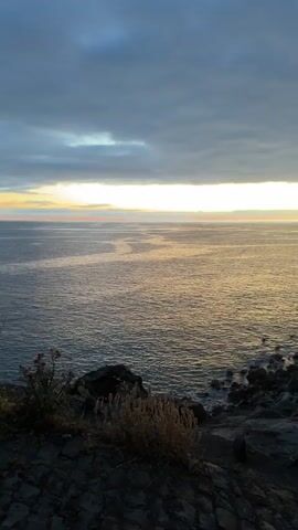 The camera pans from left to right, revealing a seascape at dusk. The sea is a shimmering gold under a cloudy sky with a streak of light on the horizon. A rocky cliff face dominates the right side of the frame, with cacti growing along the coastline.