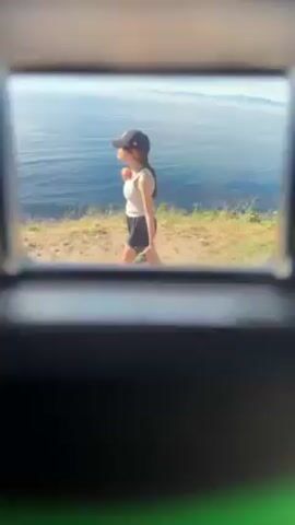 A handheld view through a dark, rectangular opening shows a young woman with dark hair, wearing a white tank top, black shorts, and a black baseball cap. She's walking along a grassy trail on a cliffside overlooking the ocean. The ocean is blue with whitecaps, and the sky is blue with some clouds. The woman is facing away from the camera, walking towards the ocean view. The foreground is mostly black, obscuring the origin of the view.