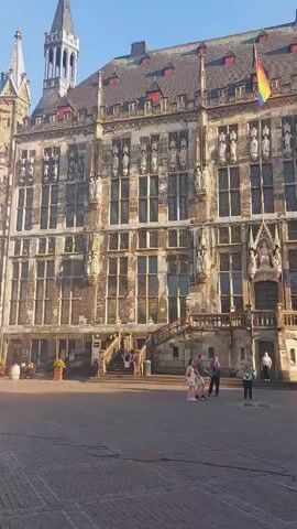 The video shows a view of the Aachen Town Hall in Germany from a public square. The building is large and ornate, with a clock tower and a rainbow flag flying from its facade. In the foreground, there is a fountain with a statue of a man on top. Several people are visible in the square, some sitting on the fountain and others walking around. The sky is clear and blue.