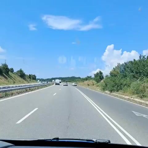 A first-person perspective video shows a vehicle traveling on a highway under a bright blue sky with scattered white clouds. The highway has multiple lanes, marked with white lines. Guardrails line the left side of the road, and green trees and vegetation are visible on the embankments. Several vehicles, including cars and a white truck, are driving ahead on the highway. The video captures the dynamic movement of the vehicle as it progresses along the road.