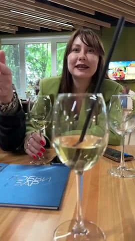 The video shows a woman sitting at a table inside a restaurant. The woman is smiling and wearing a green jacket, and she has reddish nails and a silver ring on her left hand. She holds a glass of wine with a straw in it. There is another wine glass on the table next to her, as well as a phone and a blue menu that reads "Лобби Бар". The table appears to be wooden. The background shows other people in the restaurant and a window with trees outside. A television is also visible in the background.