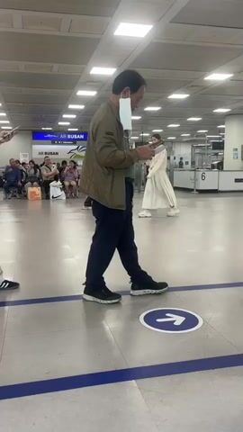 The video shows an indoor scene, possibly an airport terminal, with tiled flooring and fluorescent lighting. A man in the foreground, wearing a jacket and dark pants, walks to the right, holding some papers in his hand and a mask around his neck. He is wearing dark shoes. To his left, a person in a white t-shirt and loose-fitting light-colored pants also walks to the right. This person is wearing a backpack and is holding a mask in their right hand. Another person, a woman with glasses, walks in the same direction, carrying a green backpack and a bag with snacks. In the background, there are other people, some sitting and some walking, and what appear to be check-in counters or security gates. An Air Busan sign is visible in the distance. A blue line runs horizontally across the floor, and a circular blue and white arrow sticker indicates a direction to the right.