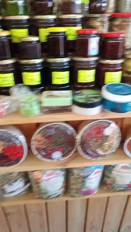 The video pans across shelves stocked with various items, primarily food products and cosmetics. The first shelf displays jars of preserves or pickled items, followed by more jars of similar contents. The second shelf features plastic containers of food items, alongside bottled liquids in red, blue, and green. The third shelf holds bags of what appear to be dried goods or teas, with some packaged food items above them. The fourth shelf presents an assortment of small jars, round containers, and bottles, likely containing spices, herbs, or cosmetics. The fifth shelf contains more jars and bottles, some with labels suggesting they are oils or tinctures, alongside what looks like soap bars and cosmetic creams. The sixth shelf is filled with various sized jars and bottles, many appearing to contain honey or syrups, along with some food products in plastic containers. The seventh shelf exhibits more jars of preserves or food items, and the final shelf is packed with bags of dry goods, possibly teas or spices. The environment appears to be a market stall or a small shop. There are no people visible in the video. The lighting is bright, suggesting an indoor setting with artificial light or natural light from an unseen source.
