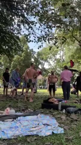 In an outdoor scene in a park, a group of approximately a dozen individuals are gathered on a grassy area dotted with fallen leaves, with trees providing a canopy overhead. One shirtless man in the center is juggling three objects, possibly hats or pads, while other members of the group are standing and watching. Some appear to be interacting with each other. The ground is covered with blankets and miscellaneous items, including bottles, clothes, and bags, suggesting a relaxed, informal setting. The overall impression is that the group is enjoying a leisurely activity in a natural environment.