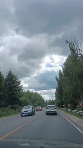 The video shows a view from the inside of a vehicle traveling on a two-lane road. The sky is cloudy with patches of blue. On either side of the road, there are trees and vegetation. There are several other vehicles on the road. In the foreground, the dashboard of the vehicle is visible. Red road markers with white arrows are visible on the right side of the road, indicating a curve. A bus stop shelter is visible on the left side of the road at one point. A dark-colored SUV passes the camera's vehicle.