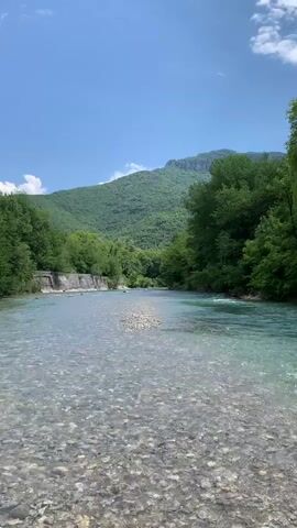 The video shows a serene river scene on a bright, sunny day. The water is clear and shallow, revealing a bed of rounded pebbles. The river flows smoothly with a gentle current. Lush green trees line both sides of the riverbank. A mountain covered in green vegetation is visible in the background under a clear blue sky with a few scattered white clouds.