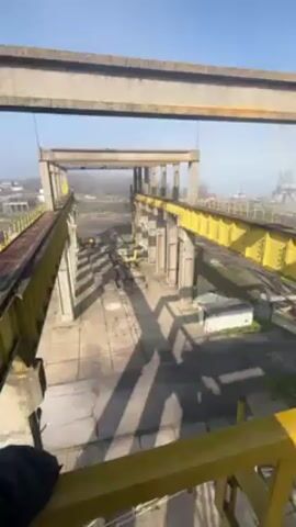 The video shows a high-angle view from atop a large, weathered concrete and steel structure painted in grey and yellow. The camera pans across a construction or industrial site with large gantry cranes in the background, near a body of water. A thick bundle of black cables hangs down, swaying slightly as the camera moves along a yellow walkway. The walkway is made of metal with a checkered texture. The sky is clear and blue.