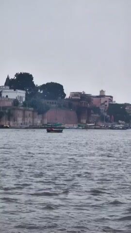 The video opens with a wide shot of a body of water, possibly a river, with moderate waves. In the background, a city skyline with traditional architecture is visible. Several buildings, some appearing old and made of stone, line the waterfront. A large wall or embankment separates the water from the city. A few boats are scattered across the water. One prominent boat in the foreground, with a green canopy, appears to be carrying passengers. The sky is overcast, suggesting a cloudy day. The video pans slightly to the right, revealing more of the city and its structures. The overall impression is of a bustling waterfront in an ancient city.