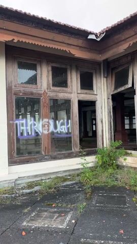 The video shows the exterior of an abandoned building. The building has wooden frames around large windows. Some of the windows are broken, while others have the word "THROUGH" spray-painted in a light purple color. The building's exterior is worn and weathered, with signs of decay and neglect. There's vegetation growing near the building's foundation. The paving stones in front of the building are cracked and covered with moss. The overall environment is one of abandonment and disrepair.