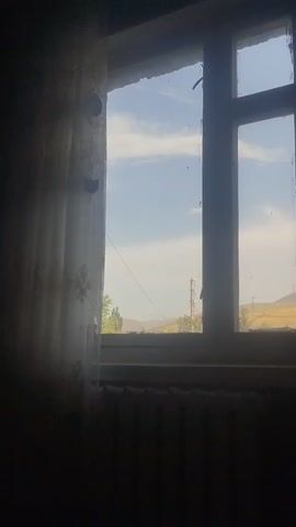 The video shows a view from a window. The window has a white frame and is divided into multiple panes by white mullions. A sheer white curtain with a floral pattern is drawn to the left, partially obscuring the view. Outside the window, the sky is blue with scattered white clouds. In the distance, there are hills and some trees. A slender, tower-like structure, possibly a utility pole or antenna, is visible against the sky. The foreground is dark, with the bottom of the window frame and what appears to be a radiator visible. The video appears to be taken indoors, looking out.