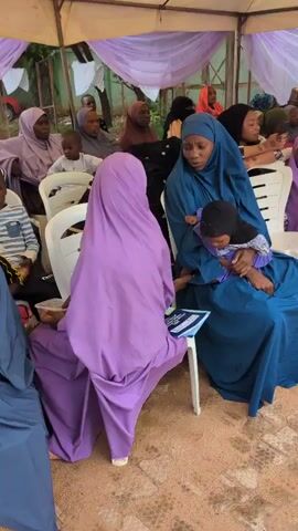 A group of people, mostly women and children wearing hijabs, are gathered under a tent. In the foreground, a woman in a blue hijab is holding a baby and talking to a child in a purple hijab. The child is sitting on a white chair, and the woman is holding a booklet or paper. Other people are seated in the background, some of whom are also wearing hijabs. The environment appears to be outdoors, with a patterned ground visible. A large draped piece of purple fabric hangs in the center of the frame.