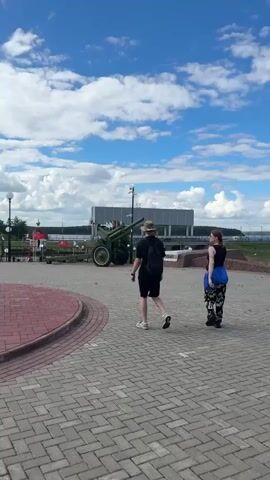 Two people are walking away from the camera on a paved walkway. The person in front is wearing a black t-shirt, black shorts, and a hat. The person behind them is wearing a black tank top, patterned pants, and has a blue bag slung over their shoulder. In the background, there is a large green cannon and a modern building. The sky is blue with white clouds. The scene appears to be an outdoor public space, possibly a park or memorial.