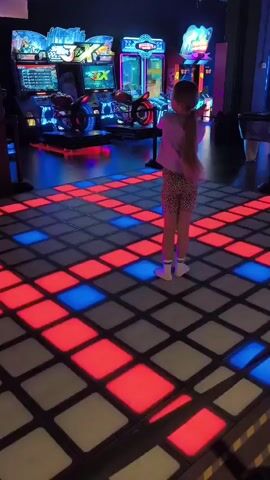 A young girl with blonde hair tied back in a ponytail, wearing a light-colored t-shirt and leopard-print shorts, dances on a lighted floor in an arcade. The floor is a grid of squares, some red and some blue, that illuminate as she steps on them. In the background, various arcade games are visible, including what appears to be a motorcycle racing game and other flashing, colorful machines. The overall environment is dimly lit, with the primary illumination coming from the arcade games and the light-up dance floor. The girl is facing away from the camera, with her arms outstretched as she moves her feet on the illuminated tiles.