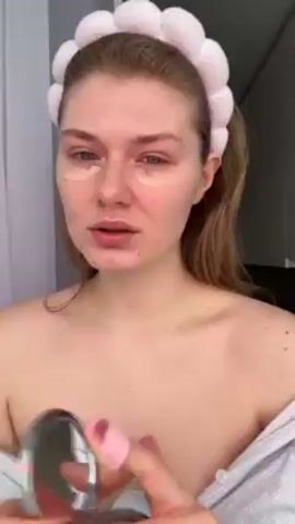 A woman in a white and light-blue striped shirt applies makeup in a bright room. She wears a fuzzy headband with white spheres. She dabs concealer under her left eye with a small pink applicator. She has light skin and reddish-blonde hair pulled back in a low ponytail. White curtains hang in the background to her left.
