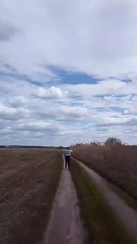 A person is jogging down a dirt path in a rural landscape. The person has their arms outstretched, almost like they're pretending to be an airplane.  To the left of the path is a vast, open field of dry, brown earth. To the right is a line of tall reeds and a few trees. The sky overhead is a mix of blue and white, with puffy clouds scattered across the expanse. The overall scene suggests a serene, open space.