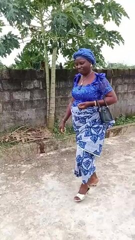 A woman in a blue and white patterned outfit and a blue headscarf walks across a paved area. She carries a black handbag. The woman turns and walks away from the camera. The environment appears to be outdoors with a wall and some vegetation in the background.