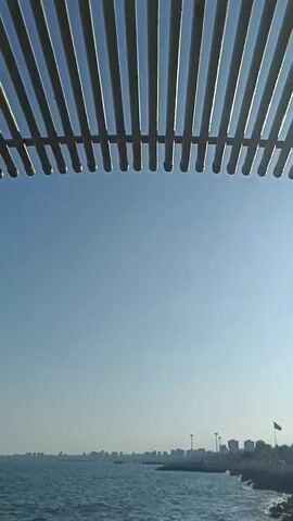 The video shows a body of water, likely the sea, with a distant cityscape visible on the horizon. The water is a deep blue and appears to be slightly choppy. There are rocks in the foreground, suggesting a shoreline or breakwater. Above the water, there is a clear blue sky. The top of the frame is partially obscured by a structure made of parallel wooden slats, which could be a pergola or a shade structure. The overall impression is of a calm, sunny day by the sea.