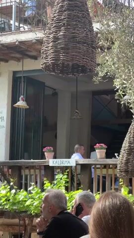The video shows an outdoor cafe or restaurant setting on a sunny day. Several people are seated at tables, some engaged in conversation, and one is on their cell phone. Above the seating area, there are large, woven, beehive-shaped decorations hanging from the ceiling. Potted pink flowers are placed on a wooden railing. The sign "KALDRMA" is visible. The cafe appears to have a balcony or upper level, as indicated by the railing and partial view of a person seated upstairs. A leafy plant or vine grows along the base of the railing. There are trees in the background.