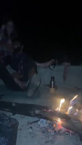 The video depicts a group of people gathered around a campfire at night. The fire provides the primary source of light. A woman wearing a plaid shirt is seated on a ledge, laughing, and a man is sitting at her feet with his arm around her. Another man is seated on the ground nearby, looking at the camera. In front of him, a person is washing something, possibly vegetables, in a blue bowl. A black trash bag is also visible on the ground. The group is engaged in a social gathering, enjoying each other's company by the campfire. The atmosphere appears relaxed and informal.