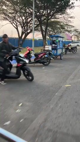 The video shows a street scene with several food carts and motorcycles. In the foreground, a blue and white food cart is attached to a motorcycle, with a person visible inside. The cart has wheels and appears to be set up for selling food. Further down the street, other food carts and motorcycles are parked. The environment appears to be an outdoor setting, possibly a roadside market or food street. The lighting suggests it might be late afternoon or early evening.
