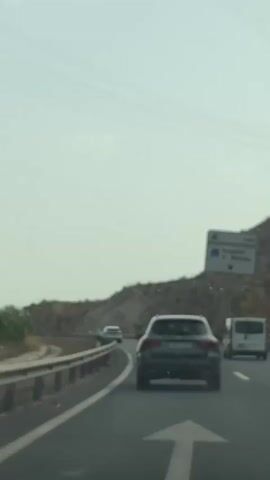 This video captures a view from inside a vehicle driving on a highway. The perspective is from the backseat or dashboard, looking forward. The primary subjects are the vehicles on the road ahead: a dark-colored SUV in the middle lane, a white van in the right lane, and a white car further ahead in the left lane. The road is a multi-lane highway with a concrete barrier on the left and a sloping, rocky hillside on the right. The sky is bright and appears hazy, suggesting daytime with possibly overcast or dusty conditions.

A prominent white road sign is visible on the right side of the highway, above the hillside. The sign contains text and symbols, including what appears to be a hospital icon and directional arrows. The text on the sign is partially legible and reads "Hospital" followed by "M. Mendez" and a directional arrow. The location context provided is El Cementerio, Spain, which helps to place the setting.

The overall activity is travel on a highway. There are no people directly visible within the frame, only the vehicles. The scene conveys a sense of movement and a journey in progress. The weather appears to be warm and bright, typical of a sunny day, but the haze in the sky might indicate pollution, dust, or high humidity. The terrain suggests a somewhat arid or mountainous region. There are no overt emotional cues conveyed by the image, but the focus on the road and the movement of vehicles implies a practical, functional activity.