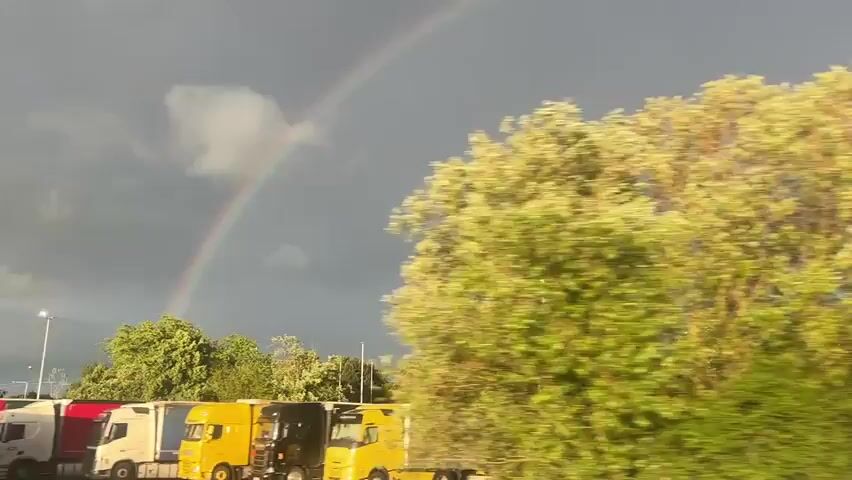 The video shows a bright rainbow against a gray sky. Below the rainbow is a line of trees and a row of parked semi-trucks. There are light poles standing in the parking lot. The video appears to be taken from a moving vehicle.