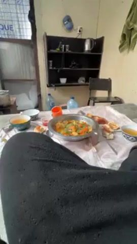 The camera shows a low-angle view of a table covered with a light-colored tablecloth, laden with food. Dominating the scene is a large metal dish filled with a vibrant orange-colored stew, garnished with green herbs, likely cilantro. Surrounding the dish are pieces of bread, small cups filled with a clear, orange liquid, possibly tea or soup, and a small container of reddish sauce. 

A person is partially visible, wearing dark, knitted pants, suggesting they are sitting or reclining on the floor around the table. The room is simply furnished with a dark-brown shelving unit, holding a kettle, glasses, and other items. Two large water bottles are placed near the shelf. A window with translucent panes is seen on the left side of the frame. A metal cooking pot is vaguely visible near the window. The overall setting is casual, possibly a home or a simple outdoor dining area.