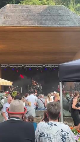 A live band performs on an outdoor stage under a wooden awning. The band consists of a drummer behind a drum set, a guitarist on the left, and a singer in the center. The stage is lit with spotlights and colored lights, and there is smoke on stage. A crowd of people watches the performance from below the stage. The audience members are diverse in age and gender, and they are seated and standing. Some are taking pictures with their phones. The environment appears to be a park or outdoor venue with sculptures visible in the foreground. The overall atmosphere is that of a casual outdoor concert.