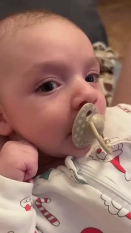 A close-up shot focuses on a baby's face as it looks towards the camera with a pacifier in its mouth. The baby is wearing a white onesie adorned with a Christmas-themed pattern featuring Santa Claus, candy canes, and Christmas trees. A beaded pacifier clip is attached to the onesie. The background is blurred but appears to be an indoor setting with a person's legs and a patterned blanket visible. The lighting suggests an ambient indoor environment.