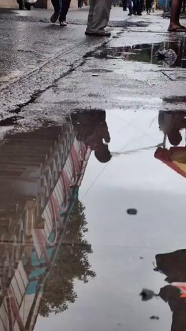 The video shows a street scene with several people walking on a wet, uneven road with puddles. In the foreground, a reflection of a person and a building is visible in a puddle. A woman in a light-colored shirt and dark pants walks from left to right, carrying a yellow object. A man wearing a maroon shirt and light-colored pants with a backpack is standing in the middle of the road, facing away from the camera. He appears to be praying or looking down. Behind him, other people are walking and a yellow auto-rickshaw is visible. The road is lined with buildings and a bridge-like structure with pink and blue accents. The sky is overcast.