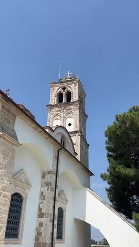 The video pans across the exterior of a white building with a bell tower. The building has stone accents around the windows and doors, and a red tiled roof. The bell tower is made of stone and has an arched opening at the top with a cross on its spire. A green tree is visible on the right side of the frame. The sky is clear and blue. The ground in front of the building is paved with cobblestones. A traffic cone is visible on the right side of the frame, near the building.