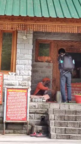 A medium shot shows two people outside a building with a green corrugated roof and stone walls. In the foreground, to the left, is a red sign with white text. To the right, a person wearing an orange robe and head covering sits on a mat on stone steps. They are facing a person in blue jeans and a backpack who is kneeling. The kneeling person is facing the entrance to the building, which is framed by ornate carved wood. Inside the entrance, a statue of a deity is visible. A bell hangs above the entrance. There are potted plants to the right of the entrance. The ground in front of the steps has a drainpipe and some red shoes. The video is shot at eye level.
