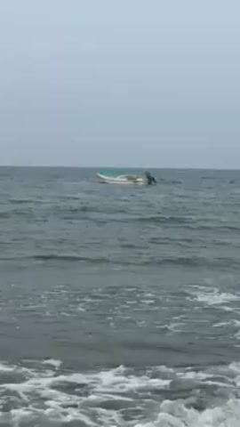 The video shows a small motorboat on the ocean, bobbing gently on the waves. The boat is white with a green stripe along the top. The water is a deep gray-blue and there are small white-capped waves. The sky is overcast and gray. The camera is positioned on the shore, capturing a view of the ocean and the boat in the distance.