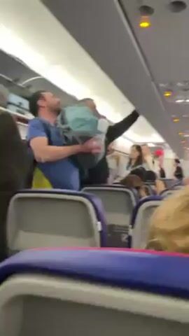 The interior of an airplane is visible. Passengers are standing in the aisle, some trying to access the overhead compartments. A man in a blue shirt struggles to lift a light blue backpack into an overhead bin. Other passengers stand nearby, some watching, some talking. The seats are blue with pink trim. The ceiling of the plane is visible, with reading lights turned on. The camera's viewpoint is from a seat, with the back of a head with blonde hair in the foreground.