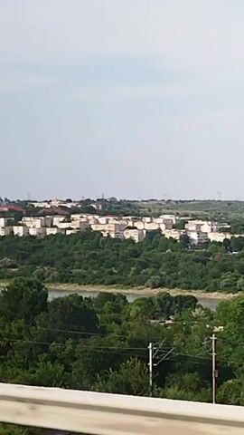 The video shows a landscape view with a river in the foreground, flanked by lush green trees and vegetation. In the middle ground, there is a town with several white apartment buildings. Further in the distance, across the river and behind the trees, is a large industrial complex with numerous wind turbines visible on the horizon. The sky is overcast with a pale blue hue. The camera appears to be moving slightly, giving a sense of a slow pan or gentle sway. There are no people or discernible actions taking place in the video. The overall impression is of a semi-urbanized area situated within a natural landscape.