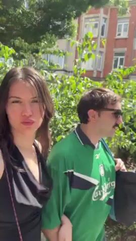 Here is a brief description of the video:

The video shows a woman and a man walking on a paved sidewalk. The woman is holding the camera and filming herself and the man. The woman has long, dark hair and is wearing a black tank top. The man has dark hair and a mustache, and he's wearing sunglasses and a green soccer jersey with light blue jeans. He has a bag slung over his shoulder. They are walking past a building and some trees. The woman makes a kissing face at the camera.