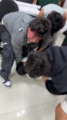 The video shows four young men struggling on the floor in a room with white tiled floors. One man is on the ground, pinned down by the other three. They are all dressed in casual clothing. A woman stands in the doorway, smiling and holding a green plastic tub. There is a chair nearby and a door labeled “Ruta de evacuacion en caso de emergencia.” The struggle appears playful, as some of the men are laughing.